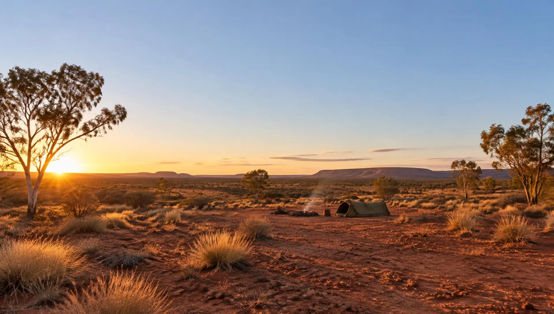 Red earth plain with eucalyptus trees at sunset and a swag beside a campfire in the distance
